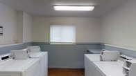 This single-floor laundry facility offers a straightforward, functional layout designed to accommodate shared or communal use. The room features a two-tone wall treatment, with a neutral upper half and a gray lower wainscot that adds a practical, easy-to-maintain finish throughout the space. A row of top-load washing machines lines one side of the room, while utility sinks with faucets are positioned opposite, providing convenient rinsing and hand-washing capabilities. Overhead, a flush-mounted fluorescent light fixture provides bright, even illumination across the entire space. A centrally positioned window with horizontal blinds allows for natural light and ventilation, helping to keep the room fresh during use. The flooring is a warm-toned hardwood, adding a touch of warmth to the otherwise utilitarian environment. The textured ceiling completes the room's no-frills, practical aesthetic. Overall, this laundry space is efficiently arranged to support multiple users simultaneously, making it well-suited for a shared residential or commercial setting.