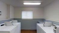 This single-floor laundry facility offers a straightforward, functional layout designed to accommodate shared or communal use. The room features a two-tone wall treatment, with a neutral upper half and a gray lower wainscot that adds a practical, easy-to-maintain finish throughout the space. A row of top-load washing machines lines one side of the room, while utility sinks with faucets are positioned opposite, providing convenient rinsing and hand-washing capabilities. Overhead, a flush-mounted fluorescent light fixture provides bright, even illumination across the entire space. A centrally positioned window with horizontal blinds allows for natural light and ventilation, helping to keep the room fresh during use. The flooring is a warm-toned hardwood, adding a touch of warmth to the otherwise utilitarian environment. The textured ceiling completes the room's no-frills, practical aesthetic. Overall, this laundry space is efficiently arranged to support multiple users simultaneously, making it well-suited for a shared residential or commercial setting.