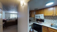 This single-story residence offers a well-organized layout with multiple rooms connected by a central corridor. The kitchen is equipped with warm honey-toned wood cabinetry providing generous storage, white appliances including an electric range, over-the-range microwave, and refrigerator, complemented by a tile mosaic backsplash and durable countertops. Wood-style flooring grounds the kitchen space. The living room features a drop tile ceiling, neutral walls, and a wide window fitted with horizontal blinds that allows natural light to filter in. A wall-mounted HVAC unit provides climate control. The bedrooms are carpeted and share a similarly neutral palette with white walls and standard windows overlooking greenery outside. Each bedroom features a closet, and the rooms vary in size, offering flexibility for different uses. The bathroom includes a mirrored medicine cabinet, a white vanity with storage, and a shower-tub combination with a curtain. Throughout the unit, drop tile and paneled ceilings are consistent features, and wall sconce lighting adds functional illumination to the hallway areas. The overall ambiance is modest, practical, and move-in ready.