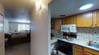 This single-story residence offers a well-organized layout with multiple rooms connected by a central corridor. The kitchen is equipped with warm honey-toned wood cabinetry providing generous storage, white appliances including an electric range, over-the-range microwave, and refrigerator, complemented by a tile mosaic backsplash and durable countertops. Wood-style flooring grounds the kitchen space. The living room features a drop tile ceiling, neutral walls, and a wide window fitted with horizontal blinds that allows natural light to filter in. A wall-mounted HVAC unit provides climate control. The bedrooms are carpeted and share a similarly neutral palette with white walls and standard windows overlooking greenery outside. Each bedroom features a closet, and the rooms vary in size, offering flexibility for different uses. The bathroom includes a mirrored medicine cabinet, a white vanity with storage, and a shower-tub combination with a curtain. Throughout the unit, drop tile and paneled ceilings are consistent features, and wall sconce lighting adds functional illumination to the hallway areas. The overall ambiance is modest, practical, and move-in ready.
