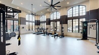 Located at 600 West State Street in Kennett Square, PA, this fitness center offers a thoughtfully designed exercise space that combines functionality with striking architectural character. The room is defined by soaring ceilings adorned with large, industrial-style ceiling fans that ensure optimal air circulation throughout the space. A row of tall, arched multi-pane windows lines the walls, flooding the interior with abundant natural light and providing views of the surrounding outdoor environment.

The two-tone wall treatment—warm beige on the lower half and deep charcoal-brown on the upper half—gives the space a modern, polished aesthetic. The light-colored flooring provides a clean, open feel underfoot, maximizing the sense of spaciousness throughout the room.

Equipment is well-curated and comprehensive. Cardio offerings include treadmills, a stationary bike, an elliptical, and a stair climber. Strength training is supported by a multi-functional cable and power rack system, a dedicated dumbbell rack, and an Octagon functional training station. Live-edge moss wall panels add a distinctive biophilic design element, complementing the mirrors that enhance depth and visibility. Entry is through sleek double glass doors, and posted fitness studio rules ensure a well-managed environment for all users.