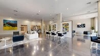 Located in the heart of downtown Sarasota at 201 S Palm Ave, the One Palm lobby is a striking, single-floor communal space that makes a bold architectural statement. Expansive polished concrete floors stretch throughout, reflecting natural light and lending the space a sleek, contemporary character. Soaring ceilings and exposed structural columns add an industrial-chic quality, while floor-to-ceiling glass windows and entry doors frame lush street-level views of palm trees and the surrounding neighborhood, flooding the interior with daylight.

The open-plan layout is thoughtfully divided into distinct functional zones. A lounge area features a white sectional sofa and accent chairs arranged around a linear fireplace framed by a dark wood surround with a mounted display above — creating an inviting focal point. Adjacent seating clusters with black leather chairs and modern white tables provide flexible gathering spaces, while a service counter and coffee bar offer added convenience for residents and guests.

Curated, large-format artwork is prominently displayed throughout, adding vibrant color and cultural character to the otherwise neutral palette of white walls and warm concrete. Potted tropical plants punctuate the space, reinforcing the Florida setting. The overall ambiance is polished, modern, and welcoming — a refined arrival experience that reflects the upscale nature of the One Palm building.