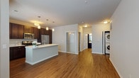 This single-floor residence showcases a thoughtfully designed open-concept layout that seamlessly connects the kitchen and living areas. The kitchen serves as the heart of the home, anchored by a spacious center island with a crisp white countertop and an undermount sink. Rich, dark-stained cabinetry extends to the upper walls, providing generous storage, while a full suite of stainless steel appliances — including a refrigerator, electric range, and over-the-range microwave — adds a sleek, contemporary finish. Stylish pendant lighting illuminates both the island and the surrounding workspace. Warm hardwood-style flooring runs throughout the main living areas, enhancing the cohesive aesthetic of the space. The open living area benefits from natural light provided by a large window, creating a bright and welcoming atmosphere. A dedicated in-unit laundry area with a stacked washer and dryer offers added convenience. The bathroom is well-appointed with dark wood cabinetry, a white countertop vanity, a large wall mirror, and a full-width glass-enclosed walk-in shower with chrome fixtures. Neutral wall tones and clean white trim throughout give the space a polished, move-in ready appearance.