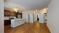This single-floor residence showcases a thoughtfully designed open-concept layout that seamlessly connects the kitchen and living areas. The kitchen serves as the heart of the home, anchored by a spacious center island with a crisp white countertop and an undermount sink. Rich, dark-stained cabinetry extends to the upper walls, providing generous storage, while a full suite of stainless steel appliances — including a refrigerator, electric range, and over-the-range microwave — adds a sleek, contemporary finish. Stylish pendant lighting illuminates both the island and the surrounding workspace. Warm hardwood-style flooring runs throughout the main living areas, enhancing the cohesive aesthetic of the space. The open living area benefits from natural light provided by a large window, creating a bright and welcoming atmosphere. A dedicated in-unit laundry area with a stacked washer and dryer offers added convenience. The bathroom is well-appointed with dark wood cabinetry, a white countertop vanity, a large wall mirror, and a full-width glass-enclosed walk-in shower with chrome fixtures. Neutral wall tones and clean white trim throughout give the space a polished, move-in ready appearance.