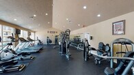 The Enclave in College Station, Texas offers a comprehensive fitness center designed to support a full range of workout routines. The single-floor facility is thoughtfully laid out, with distinct zones for cardio, strength training, and additional wellness amenities. The cardio area lines the windowed wall, where treadmills and elliptical machines face expansive floor-to-ceiling windows that overlook the outdoor pool area, providing an energizing view and ample natural light during workouts. Mounted televisions add to the convenience of the cardio zone. The strength training section is well-appointed with a variety of cable and resistance machines, including multi-function cable stations and targeted isolation equipment. A dedicated free weight area features a full dumbbell rack with a mirrored wall, allowing users to monitor their form. Rubber flooring throughout the main workout space ensures safety and durability. The facility also includes a private tanning room, equipped with a Sunvision tanning bed and accented with decorative tropical plants, offering a relaxing retreat within the fitness center. Recessed lighting illuminates the entire space, while Aggie-themed wall decor adds a nod to the local Texas A&M community. The fitness center at The Enclave provides a well-rounded amenity for residents seeking a convenient and fully equipped place to maintain an active lifestyle.