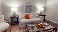 This single-floor residence offers a practical and cohesive layout across several well-defined living spaces. The galley-style kitchen is efficiently organized with gray cabinetry lining both sides, dark granite-style countertops, and black appliances including a refrigerator, electric range, and over-the-range microwave. Light wood-look flooring runs throughout the kitchen, adding brightness to the space. A peninsula with a dark countertop provides a natural transition into the adjacent dining area, which features light flooring, a round wood dining table, and a window dressed with soft blue curtains that allow natural light to filter in. A chandelier overhead adds a subtle decorative touch. The living room is finished with dark wood-look flooring, neutral gray walls, and a textured ceiling, creating a relaxed and inviting atmosphere. The bedroom continues the dark flooring theme and connects directly to the hallway, offering easy access to additional rooms. The bathroom includes a full tub-shower combination with classic white subway tile surround and a curtain rod. Throughout the unit, textured walls and ceilings, neutral tones, and consistent flooring choices contribute to a cohesive, understated aesthetic.