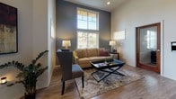 This single-floor space is defined by its notably high ceilings that lend an open, airy quality throughout. Light wood-look flooring runs continuously across the interior, unifying the various functional areas and contributing to a clean, modern aesthetic.

The entry area welcomes visitors with a pendant light fixture, a dark metal storage console adorned with artwork, and a glass-paneled front door that draws in natural light. The space opens into a generously sized common area that accommodates multiple distinct zones.

A lounge area features a neutral-toned sofa with accent pillows, a glass-top coffee table, and tall windows that rise toward the ceiling, flooding the space with abundant natural light and outdoor views. Dark charcoal accent walls provide a grounded contrast to the lighter surrounding walls, enhancing the cozy atmosphere of this seating zone.

Adjacent to the lounge, a casual dining area includes a round table with modern chairs, a wall-mounted flat-screen television, and an ornate entertainment console with a built-in electric fireplace below. A wall-mounted mini-split unit ensures climate comfort. Interior windows connect this area visually to neighboring spaces, maintaining an open and connected feel throughout the layout.