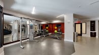 Located at 10914 Georgia Ave in Silver Spring, Maryland, this fitness center offers a generously sized, single-floor exercise space designed to support a full range of workout routines. The layout is open and thoughtfully organized, with distinct zones dedicated to cardio equipment, strength machines, and free weights. Treadmills, ellipticals, and stationary bikes are positioned along one end of the room, complemented by a bold, motivational typography mural that spans the full length of the accent wall, creating an energizing atmosphere. A vibrant red accent wall anchors the free weights area, where a multi-rack dumbbell station, stability balls, and a cable resistance machine are neatly arranged. Large floor-to-ceiling mirrors line the walls, enhancing the sense of openness and allowing users to monitor their form. Weight benches and additional resistance machines occupy the central floor space. The polished concrete flooring is durable and easy to maintain, while recessed linear lighting ensures bright, even illumination throughout. Stainless steel waste receptacles and hand sanitizer dispensers are conveniently placed around the room. Glass-panel entry doors at the front of the space allow natural light to filter in, contributing to an inviting and well-lit environment suited for focused, effective training sessions.
