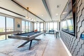 Located in Dallas, TX, this contemporary sky lounge is a thoughtfully designed amenity space that seamlessly blends entertainment and relaxation. The open single-floor layout is anchored by a sleek, modern billiards table positioned atop a marbled area rug, illuminated by a striking brass pendant light fixture that adds a warm, sophisticated glow. Floor-to-ceiling windows and large glass doors flood the space with abundant natural light while framing expansive elevated views of the surrounding Dallas skyline, treetops, and neighboring buildings. A textured hexagonal tile accent wall provides a bold focal point, complemented by a large mounted flat-screen display — ideal for entertainment gatherings. On one side, a galley-style wet bar features dual dark cabinetry runs with stone countertops, a geometric white tile backsplash, an undermount sink, and integrated appliances, providing a fully functional serving area. The lounge area near the terrace access offers comfortable seating with linen drapery and direct access to an outdoor terrace strung with ambient bistro lighting. Large-format tile flooring ties the entire space together, lending a clean, polished aesthetic throughout. This sky lounge delivers a refined, upscale environment suited for socializing and entertainment.