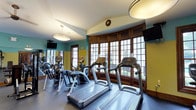 This single-floor fitness center offers a thoughtfully designed workout environment that blends functionality with classic architectural charm. The space is anchored by a row of modern treadmills and elliptical cross-trainers positioned to take full advantage of the expansive wood-trimmed grid windows that line the far wall. These large windows flood the room with natural light and provide views to the outdoors, creating an open, energizing atmosphere for exercise. Additional cardio and strength equipment is arranged along the perimeter, maximizing usable floor space without feeling cramped. The vaulted ceiling rises at an angle above the room, adding a sense of spaciousness and height. Overhead, elegant drum-style pendant light fixtures complement the room's architectural character, while wall-mounted flat-screen televisions offer entertainment during workouts. The walls feature a warm combination of teal and yellow-green tones, adding a vibrant yet inviting energy to the space. Rich wood baseboards and window frames throughout provide a refined, classic aesthetic that distinguishes this fitness center from a typical gym environment. The durable, dark-toned flooring is well-suited for high-traffic use. Overall, the space strikes a balance between practical fitness amenities and an upscale, welcoming design sensibility.