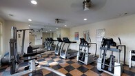 Located at 300 Cosgrove Avenue in Chapel Hill, North Carolina, this fitness center offers a well-appointed, single-floor workout facility designed to support a full range of exercise routines. The space features a distinctive checkered floor in black and tan tones, lending the room a bold, clean aesthetic that complements its functional purpose.

The cardio area is prominently featured, housing a row of treadmills and elliptical machines positioned for ease of use and proper spacing between equipment. A large wall mirror runs along one side of the room, visually expanding the space and allowing users to monitor their form during workouts.

Ceiling fans and recessed lighting provide consistent, comfortable illumination throughout, ensuring the space remains bright and well-ventilated during use. Instructional workout charts and fitness reference posters are mounted on the walls, offering guidance for exercise routines and muscle group targeting.

Additional strength and conditioning equipment is visible along the perimeter of the room, rounding out the facility's offerings. The overall ambiance is clean, organized, and purposefully designed, creating a motivating environment suitable for both casual and dedicated fitness enthusiasts.