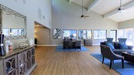 This single-floor space makes a strong first impression with its dramatic vaulted ceilings accented by exposed beam detailing and a ceiling fan that enhances the sense of airiness throughout. The open-plan layout is anchored by a central workspace featuring a large desk, upholstered seating, and a blue area rug that defines the functional zone. Warm hardwood-style flooring extends throughout, complementing the cool grey-toned walls and crisp white trim. A row of large windows along the back wall floods the interior with abundant natural light, offering views to the outdoors and creating a bright, inviting atmosphere. Decorative framed artwork adds personality to the walls, while a stylish console table near the entrance provides a welcoming focal point. An adjoining room is visible through an open archway, suggesting additional functional areas within the space. The overall ambiance is professional yet comfortable, balancing open-concept design with purposeful furnishings. The generous ceiling height and expansive floor area contribute to a feeling of openness that is both functional and visually impressive, making it well-suited for a leasing office, reception area, or similar professional environment.