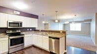 This single-floor residence offers an open-concept layout that connects the kitchen and living areas in a cohesive, functional design. The kitchen is a standout feature, showcasing white cabinetry that extends to the ceiling, dark granite countertops, and stainless steel appliances including a range, dishwasher, and built-in microwave. A peninsula with a double basin sink serves as a natural dividing point between the kitchen and the rest of the living space, providing additional prep area and casual seating potential. Pendant lighting with brushed nickel fixtures hangs above the peninsula, adding both task lighting and a modern aesthetic touch. The rich, dark hardwood flooring in the kitchen transitions into carpeted flooring in the adjacent living area, offering a warm contrast. The living space is brightened by natural light streaming through large windows, and a ceiling fan helps maintain comfort throughout. Dark accent walls provide depth and visual interest against the lighter gray tones that dominate the walls. The overall ambiance of the space is modern, comfortable, and well-appointed, making it both practical for everyday living and inviting for entertaining.