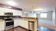 This single-floor residence offers an open-concept layout that connects the kitchen and living areas in a cohesive, functional design. The kitchen is a standout feature, showcasing white cabinetry that extends to the ceiling, dark granite countertops, and stainless steel appliances including a range, dishwasher, and built-in microwave. A peninsula with a double basin sink serves as a natural dividing point between the kitchen and the rest of the living space, providing additional prep area and casual seating potential. Pendant lighting with brushed nickel fixtures hangs above the peninsula, adding both task lighting and a modern aesthetic touch. The rich, dark hardwood flooring in the kitchen transitions into carpeted flooring in the adjacent living area, offering a warm contrast. The living space is brightened by natural light streaming through large windows, and a ceiling fan helps maintain comfort throughout. Dark accent walls provide depth and visual interest against the lighter gray tones that dominate the walls. The overall ambiance of the space is modern, comfortable, and well-appointed, making it both practical for everyday living and inviting for entertaining.