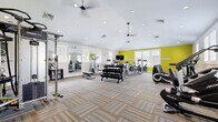 Located in Hampton, Virginia, this fitness center offers a generously sized, single-floor open layout designed to accommodate a comprehensive range of workout activities. The space is immediately defined by its striking yellow accent walls, which contrast boldly against the clean white walls and vaulted ceiling, creating an energetic and motivating atmosphere. Recessed lighting throughout the ceiling, complemented by multiple ceiling fans, ensures the space remains bright and well-ventilated during workouts.

The flooring consists of patterned, multi-tone carpet tiles that add both visual texture and practical cushioning underfoot. Large windows along multiple walls flood the interior with abundant natural light, enhancing the open and airy feel of the space.

Equipment is thoughtfully arranged throughout the room to maximize usability. The cardio zone features treadmills, elliptical trainers, stair climbers, and stationary bikes. The strength training area includes a multi-rack dumbbell station with a full range of free weights, a weight bench, and multiple cable and selectorized resistance machines. A dedicated functional training zone offers stability balls, medicine balls, and a cable pull system. Floor-to-ceiling mirrors line one wall, expanding the visual depth of the room and allowing users to monitor their form. A wall-mounted television serves as an entertainment focal point.