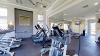 This fitness center offers a thoughtfully designed workout environment that balances functionality with refined aesthetics. The space features a distinctive coffered ceiling with clean white beams, lending an architectural elegance uncommon in typical gym settings. Large windows along the walls invite abundant natural light, creating a bright and energizing atmosphere ideal for exercise.

The facility is well-stocked with a variety of cardio equipment, including treadmills, elliptical trainers, and stationary bikes from quality brands such as True Fitness. A dedicated free weights area provides a dumbbell rack alongside benches, catering to strength training needs. Floor-to-ceiling mirrors line one wall, visually expanding the space and allowing users to monitor their form during workouts. Mounted flat-screen televisions are positioned for easy viewing from the cardio machines, enhancing the overall exercise experience.

The flooring consists of durable, dark rubberized material designed for both safety and performance. Soft neutral tones in gray and white throughout the walls and trim contribute to a clean, modern aesthetic. A built-in cabinet along one wall adds a practical storage element, while a piece of wall art adds a welcoming, polished touch. Overall, this fitness center combines upscale design with comprehensive amenities.