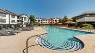 Located at 211 North Ave in Athens, Georgia, this outdoor amenity space offers a resort-style experience designed for relaxation and recreation. The centerpiece is a generously sized swimming pool with a distinctive freeform shape, featuring gradual entry steps and a shallow end clearly marked for safety. The crystal-clear water is framed by a spacious concrete sundeck outfitted with numerous chaise lounges and shaded umbrella stations, providing ample room for sunbathing and socializing.

Adjacent to the main pool area, a secondary courtyard features stylish modular lounge furniture, additional seating arrangements under striped market umbrellas, and a bistro-style table setting surrounded by meticulously maintained hedgerows and mature trees. The lush landscaping throughout creates a serene, park-like atmosphere.

A dedicated recreational zone includes a full-size basketball hoop and a large-format Connect Four game set on artificial turf, offering entertainment options for a variety of interests. The entire pool area is enclosed by a sleek black iron fence, ensuring privacy and security, with posted hours of operation from 10:00 AM to 9:00 PM.

A hip-roofed pavilion structure anchors one end of the pool deck, providing shade and additional gathering space. The surrounding multi-story residential buildings with white horizontal siding and balconies provide a warm residential backdrop to this well-appointed outdoor retreat.