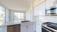 This single-floor residence showcases a clean, contemporary design throughout. The open-plan living and kitchen area serves as the heart of the space, where light wood-tone flooring flows seamlessly underfoot. A dramatic wall of floor-to-ceiling windows and glass sliding doors floods the living area with natural light while offering expansive outdoor views of the surrounding cityscape and greenery, creating a bright and airy atmosphere.

The kitchen is thoughtfully designed with high-gloss white cabinetry extending to the ceiling, providing generous storage. White quartz countertops and a classic subway tile backsplash complement the suite of stainless steel appliances, including a gas range, over-the-range microwave, and dishwasher. A peninsula with an integrated sink adds functionality and defines the transition between the kitchen and living areas.

The bedroom continues the light-filled theme with a ceiling fan, sliding glass door to a private balcony, and crisp white walls. The bathroom features warm wood-look flooring, a tiled shower-tub combination, a sleek vanity with an illuminated mirror, and clean white walls that maintain the unit's cohesive modern aesthetic throughout.