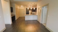 This single-floor residence showcases an open-concept layout that seamlessly connects the living area to the kitchen, creating a spacious and cohesive flow throughout. Rich, dark hardwood flooring spans the entire main area, grounding the space with warmth and continuity.

The kitchen serves as the centerpiece, featuring dark cabinetry that contrasts elegantly with light granite countertops. A peninsula with a built-in sink offers both additional workspace and a natural divider between the kitchen and living area. Stainless steel appliances, including a full-size refrigerator and microwave, complement the modern aesthetic. Above the peninsula, a set of pendant lights with elongated shades provides focused task lighting, while a track lighting system overhead ensures the kitchen remains bright and functional.

Multiple doors visible along the hallway suggest well-distributed private rooms and closet storage, including what appears to be a louvered closet door. The neutral wall tones throughout keep the space feeling clean and versatile. Overall, the unit delivers a contemporary, well-appointed interior with a practical and polished design.