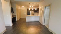 This single-floor residence showcases an open-concept layout that seamlessly connects the living area to the kitchen, creating a spacious and cohesive flow throughout. Rich, dark hardwood flooring spans the entire main area, grounding the space with warmth and continuity.

The kitchen serves as the centerpiece, featuring dark cabinetry that contrasts elegantly with light granite countertops. A peninsula with a built-in sink offers both additional workspace and a natural divider between the kitchen and living area. Stainless steel appliances, including a full-size refrigerator and microwave, complement the modern aesthetic. Above the peninsula, a set of pendant lights with elongated shades provides focused task lighting, while a track lighting system overhead ensures the kitchen remains bright and functional.

Multiple doors visible along the hallway suggest well-distributed private rooms and closet storage, including what appears to be a louvered closet door. The neutral wall tones throughout keep the space feeling clean and versatile. Overall, the unit delivers a contemporary, well-appointed interior with a practical and polished design.