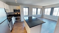 This single-floor space showcases an open-concept layout that seamlessly connects the kitchen and living area, creating a cohesive and functional environment. The kitchen is outfitted with white cabinetry that extends to the ceiling, providing ample storage, and is complemented by dark stone countertops that offer a striking contrast. A central island with matching dark countertop serves as both a prep space and a natural dividing point between the kitchen and the adjoining living area. Stainless steel appliances, including a range, oven, and built-in microwave, add a polished, contemporary touch. The flooring throughout is warm-toned hardwood, contributing to a cohesive and inviting atmosphere. Large windows along the far wall flood the living area with natural light and frame expansive city views, making the space feel open and elevated. The neutral grey wall tones and textured ceiling complement the overall modern aesthetic. The open flow between the kitchen and living areas makes this space well-suited for both everyday living and entertaining.