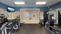 This fitness center offers a thorough and well-organized workout environment designed to accommodate a wide range of exercise routines. The space is finished with deep blue-gray walls and durable rubber flooring, creating a focused and energizing atmosphere throughout.

The cardio area features multiple treadmills and an elliptical machine, each positioned to take advantage of the abundant natural light streaming through large, multi-pane windows that frame views of mature trees and lush greenery outside. A wall-mounted television is conveniently positioned for entertainment during cardio sessions.

The strength training section is generously appointed with a variety of cable machines, weight stations, and selectorized equipment, suitable for both upper and lower body workouts. A full rack of dumbbells lines the wall near the windows, accompanied by an adjustable weight bench. Floor-to-ceiling mirrors span one wall, enhancing the sense of space and allowing for proper form checks during exercise.

A secondary alcove area provides additional functional space and includes a water fountain for convenience. Posted fitness center rules and a tiled entryway transition cleanly into the main workout floor. Overhead fluorescent lighting supplements the natural light, ensuring the space remains well-lit at all hours.