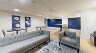 This thoughtfully designed single-floor communal space offers a versatile and inviting environment across a comfortable open-plan layout. The interior is defined by a striking navy blue and crisp white color scheme, carried consistently throughout via bold accent walls, coordinating artwork, and carefully selected furnishings.

The space is divided into several functional zones that flow seamlessly into one another. A lounge area anchors one end of the room, featuring a tufted grey sofa and matching armchair arranged atop a patterned area rug, complemented by a wall-mounted flat-screen television set against a deep navy feature wall. An elegant arched architectural detail frames the transition between areas, adding a refined structural element.

Adjacent to the lounge, a dining and social zone offers multiple black-top tables paired with industrial-style metal chairs, ideal for casual seating or collaborative use. Decorative floral canvas artwork in blue tones adorns the white walls, reinforcing the cohesive design.

A kitchenette area at the far end includes a long counter with bar-height seating, teal subway tile backsplash, vibrant orange cabinetry, and built-in appliances, providing practical amenity for gatherings. Throughout the space, light hardwood flooring, recessed circular ceiling lights, and linear LED fixtures ensure bright, even illumination, creating a welcoming and functional atmosphere.