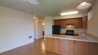 This single-floor space offers a practical and welcoming layout centered around an open-concept design that connects the kitchen and living areas seamlessly. The kitchen is well-equipped with warm medium-toned wood cabinetry, black appliances including a refrigerator and gas range, and a peninsula breakfast bar topped with a light granite-style countertop — ideal for casual dining or meal prep. An overhead range hood and under-cabinet lighting provide functional illumination in the cooking area. The flooring throughout is a warm hardwood-style material that adds continuity and a sense of warmth across the space. The living area benefits from a ceiling fan, contributing to comfortable year-round airflow. Neutral-toned walls keep the interior bright and versatile, allowing for a variety of décor styles. A hallway visible from the main living area leads to additional rooms, suggesting a well-organized floor plan. A wall vent ensures climate control throughout. The overall ambiance is clean, functional, and inviting, making it a practical space suited for everyday living.