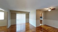 This single-floor space offers a clean, functional layout with a warm and inviting ambiance throughout. The living area is a standout feature, showcasing richly polished hardwood floors that reflect natural light streaming in through a large sliding glass door dressed with vertical blinds. The two-tone wall treatment — warm taupe on the upper half and crisp white wainscoting below — adds a classic, structured aesthetic to the space.

A partial wall thoughtfully separates the living area from the adjoining kitchen, maintaining an open feel while defining each zone. The kitchen is visible through a passthrough opening and features warm honey-toned wood cabinetry, a tile backsplash with a subtle pattern, and a sink positioned beneath upper cabinets. The flooring transitions to tile in the kitchen, providing a practical surface in the cooking area.

The overall layout is straightforward and well-proportioned, making efficient use of the available space. Natural light plays a key role in the ambiance, brightening the interior and highlighting the warm tones of the hardwood floors. The neutral color palette throughout creates a versatile backdrop suitable for a variety of interior styles. The space presents a comfortable, move-in-ready environment with practical finishes and a cohesive design sensibility.