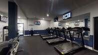 Located at 4460 Mountain Laurel Rd in Hilliard, Ohio, this fitness center offers a well-appointed, single-floor workout facility designed to support a comprehensive exercise routine. The space features a distinctive two-tone color scheme with crisp white upper walls and a bold navy blue accent band running along the lower portion, giving the gym a clean and professional aesthetic. Dark, commercial-grade rubber flooring extends throughout, providing both durability and comfort underfoot.

The cardio section is generously equipped with a row of treadmills, elliptical trainers, and stationary bikes, each positioned for easy use. Multiple wall-mounted flat-screen televisions are strategically placed throughout the space, allowing members to stay entertained during workouts. The strength training area features a variety of resistance machines and cable systems, catering to a range of fitness goals.

A standout feature of the space is a large motivational mural wall displaying words such as "commitment," "care deeply," "dedication," and "inspire," adding an energizing and uplifting atmosphere to the environment. Full-length mirrors line portions of the walls, enhancing the sense of openness. Recessed lighting keeps the facility bright and welcoming, while ceiling fans contribute to ventilation and comfort. The layout flows efficiently, offering ample room to move between equipment areas.