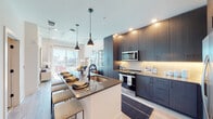 This contemporary urban residence showcases thoughtful design and premium finishes across a single-floor layout. The open-concept kitchen is a standout feature, outfitted with dark, floor-to-ceiling cabinetry, stainless steel appliances including a French door refrigerator, a tile backsplash with under-cabinet lighting, and a spacious center island with a quartz-style countertop, undermount sink, and seating — ideal for both cooking and casual dining. Stylish pendant lighting anchors the space with a modern aesthetic.

The living area is bright and airy, benefiting from expansive floor-to-ceiling sliding glass doors that open to a private balcony with striking panoramic city skyline views. Exposed concrete ceiling elements and structural columns add an industrial character that complements the contemporary furnishings.

The bedroom offers generous proportions with soaring ceilings, light wood-style flooring, and large windows that frame sweeping cityscape views, creating a serene yet connected retreat.

The bathroom features a double-sink vanity with dark cabinetry and matte black hardware, a wide frameless mirror, and a walk-in glass-enclosed shower adorned with full-height marble-style tile. Light wood flooring runs consistently throughout, unifying the entire residence with a clean, cohesive feel.