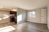 This well-appointed single-floor residence offers a clean, modern aesthetic defined by neutral gray walls, bright white trim, and light hardwood-style flooring that flows seamlessly throughout the space. The open-concept living area is generously proportioned, bathed in natural light streaming through a large window that frames views of mature trees outside. Flush-mount ceiling fixtures provide warm, consistent overhead lighting across the space.

The kitchen is positioned adjacent to the living area and features rich cherry wood cabinetry paired with dark countertops and stainless steel appliances, including a full-size refrigerator, creating a striking contrast against the lighter tones of the surrounding space. The layout allows for an easy connection between cooking and living areas.

A full bathroom is finished in crisp white tiling, complete with a tub-shower combination, a window with a privacy blind that brings in natural light, a white vanity, and hardwood-style flooring that mirrors the rest of the unit. Additional rooms include a bedroom or flexible living space with softly tinted walls and a paneled white door, offering a calm and private retreat. A staircase with dark wood treads and white painted risers, fitted with a handrail, is also visible, adding a classic structural detail to the interior.