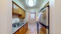 This single-floor unit offers a practical and straightforward layout across several well-defined spaces. The galley-style kitchen is efficiently designed, featuring warm wood cabinetry, granite-style laminate countertops, and stainless steel appliances including an electric range with overhead ventilation, a full-size refrigerator, and a double-basin sink. Wood-look flooring runs through the kitchen, which connects to an adjacent area through an open doorway, allowing natural light to filter in from a window overlooking surrounding trees.

The living area is generously sized, finished with neutral carpet and equipped with a ceiling fan and baseboard heating. Dual windows along one wall bring in natural light and outdoor views of mature trees. A wall-mounted air conditioning unit provides supplemental climate control.

The bedroom areas are carpeted and offer a quiet, neutral atmosphere. Sliding wood-panel closet doors provide ample storage, and one bedroom includes an additional walk-in closet visible through the doorway. The full bathroom features wood-look flooring, a single-vanity with cabinet storage, a toilet, and a tub-shower combination with clean white surrounds and a grab bar for added convenience. Overall, the space presents a clean, functional interior with consistent finishes throughout.