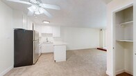 This single-floor residence offers a practical and comfortable layout across a generously sized floor plan. The open-concept kitchen and living area serves as the heart of the home, featuring white cabinetry, a peninsula counter, stainless steel appliances including a refrigerator and gas range, and tile flooring that transitions seamlessly into plush wall-to-wall carpeting in the living space. A ceiling fan overhead provides both comfort and light, while vertical blinds on a large window allow natural light to filter in. A distinctive low wooden platform near the window adds a subtle architectural accent to the living area. The bedroom is a well-proportioned retreat finished with soft carpeting, a sun-filled window with blinds, and an expansive triple-panel sliding closet for generous storage. A separate walk-in style closet area off the bedroom provides additional organization space. The bathroom is clean and functional, featuring tile flooring, a full tub-shower combo with sliding glass doors, and a vanity with storage. A hallway connects the rooms, and the neutral, light color palette throughout creates a bright and cohesive ambiance suited for comfortable everyday living.
