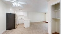This single-floor residence offers a practical and comfortable layout across a generously sized floor plan. The open-concept kitchen and living area serves as the heart of the home, featuring white cabinetry, a peninsula counter, stainless steel appliances including a refrigerator and gas range, and tile flooring that transitions seamlessly into plush wall-to-wall carpeting in the living space. A ceiling fan overhead provides both comfort and light, while vertical blinds on a large window allow natural light to filter in. A distinctive low wooden platform near the window adds a subtle architectural accent to the living area. The bedroom is a well-proportioned retreat finished with soft carpeting, a sun-filled window with blinds, and an expansive triple-panel sliding closet for generous storage. A separate walk-in style closet area off the bedroom provides additional organization space. The bathroom is clean and functional, featuring tile flooring, a full tub-shower combo with sliding glass doors, and a vanity with storage. A hallway connects the rooms, and the neutral, light color palette throughout creates a bright and cohesive ambiance suited for comfortable everyday living.
