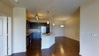 This single-floor space showcases a well-designed open-concept layout that seamlessly connects the kitchen and living areas. The kitchen is a standout feature, outfitted with dark espresso cabinetry that extends to the upper walls, providing generous storage throughout. Stainless steel appliances, including a full-sized refrigerator and built-in microwave, complement the kitchen's modern aesthetic. The countertops feature a neutral stone finish, adding a refined touch to the workspace.

A curved kitchen island serves as the centerpiece of the space, equipped with a double basin sink and offering additional seating and prep space. Three elegant pendant lights hang above the island at varying heights, enhancing both functionality and visual interest.

Rich, dark hardwood floors extend throughout the entire space, creating a warm and cohesive feel. Crown molding lines the upper walls of the living area, adding an architectural detail that elevates the overall design. A hallway connects additional rooms, offering a sense of privacy from the main living space. The neutral wall tones and warm ambient lighting contribute to an inviting, comfortable atmosphere throughout.