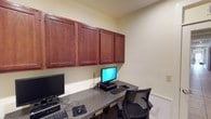 Located at 147 Links Dr in Canton, Mississippi, this single-story space is designed to support a focused and functional work environment. The interior reflects a clean, professional aesthetic with neutral-toned walls that create a calm and productive atmosphere throughout.

A standout feature of the space is the dedicated computer workstation area, which is outfitted with a long granite-finish built-in desk that comfortably accommodates multiple monitors and workstations side by side. Above the desk, a row of rich, dark wood upper cabinets with raised panel doors spans the full width of the wall, providing generous enclosed storage while adding a polished, finished look to the room. The cabinetry's deep mahogany tone contrasts attractively with the light walls, lending warmth to the otherwise streamlined office setting.

The workstation area is equipped with ergonomic mesh office chairs, supporting comfort during extended use. The practical layout ensures that equipment, storage, and seating are efficiently organized within the space. Overall, this single-floor property offers a well-appointed, detail-oriented interior suited for administrative or professional office use.