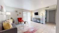 This single-floor residence offers a thoughtfully arranged layout that balances comfort and functionality throughout. The living room serves as the heart of the home, featuring large sliding glass doors dressed with vertical blinds that flood the space with natural light and provide outdoor views. Light ceramic tile flooring extends throughout, lending a cohesive and easy-to-maintain aesthetic across all rooms. Retro-inspired starburst wall décor and modern accent lighting add character to the living area, which comfortably accommodates a sofa, accent chair, and coffee table arrangement.

The galley-style kitchen is efficiently designed with gray cabinetry, light countertops, a stainless steel sink, and white appliances including a range, dishwasher, and refrigerator. A pass-through opening connects the kitchen to a dedicated dining area, which features a modern industrial-style table with bench seating and is brightened by a nearby window.

The bedroom is generously proportioned, with vaulted-style angled ceilings that enhance the sense of openness. A window provides natural light, and the neutral color palette creates a calm, restful atmosphere. The bathroom is clean and functional, highlighted by a gray vanity with ample cabinet storage, a framed mirror, and a built-in linen closet with open shelving — ideal for organized storage.