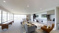 This contemporary single-floor space is designed with an open-plan layout that seamlessly integrates multiple functional areas into one cohesive, inviting environment. The expansive floor-to-ceiling windows along the far wall flood the interior with abundant natural light while offering sweeping panoramic views of the surrounding landscape, creating a bright and airy ambiance throughout.

The lounge area serves as a central gathering point, furnished with plush gray sofas and accent chairs arranged around a sleek coffee table, all set atop a stylishly patterned area rug that adds texture and warmth to the space. Gold decorative accents provide an elegant finishing touch to the seating arrangement.

Adjacent to the lounge, a fully equipped kitchen is integrated into the space, featuring modern stainless steel appliances including a refrigerator, oven, and countertop workspace with a sink. Dark cabinetry provides a sophisticated contrast to the light countertops and white walls. A large wall-mounted flat-screen television anchors the media area nearby, making this space ideal for both relaxed gatherings and entertainment.

A dining area positioned near the windows takes full advantage of the natural light and views. The smooth white ceilings with recessed lighting ensure consistent, well-distributed illumination throughout, complementing the modern and polished aesthetic of the entire space.