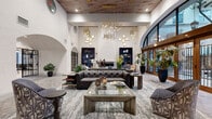 This expansive single-floor lounge space showcases a masterful blend of classical architecture and contemporary interior design. The most immediately striking feature is the soaring ceiling height, accented by warm wood ceiling panels and an elaborate cascading installation of gold brass pendant lights that descend at varying lengths, creating a dynamic, sculptural focal point throughout the space.

The architectural design is defined by graceful arched openings, including a grand floor-to-ceiling arched window wall framed in rich wood, allowing abundant natural light to flood the interior while offering views of mature trees outside. Decorative wrought-iron gated archways provide additional visual interest and define transitions between zones within the space.

The lounge is organized around multiple seating arrangements, anchored by a tufted leather Chesterfield sofa, upholstered accent chairs, and layered area rugs atop marble-look tile flooring. A sleek, modern linear fireplace serves as a refined focal point along one wall, flanked by blue diamond-patterned display niches showcasing curated art objects.

Lush potted plants are thoughtfully positioned throughout, adding organic warmth and contrast to the polished, neutral palette of whites, grays, and gold tones. The overall ambiance is refined, welcoming, and design-forward.