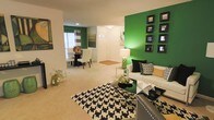 This single-floor home makes a striking first impression with its bold design choices and open, flowing layout. The living area serves as the centerpiece of the space, anchored by a vivid green accent wall adorned with a grid of dark-framed mirrors that add depth and visual interest. Plush carpeting in a neutral tone runs throughout, providing a soft contrast to the energetic color palette. The living room is furnished with a sleek white sofa dressed in patterned throw pillows, complemented by a black and white geometric area rug that adds a modern, graphic edge. A glass coffee table with decorative accents ties the space together with a polished finish. The open layout connects seamlessly to the dining area, which features a classic round table with white upholstered chairs, positioned near a window dressed with striped curtains that allow natural light to filter in. A secondary green accent wall in the dining area creates a cohesive visual thread throughout the home. The entryway is welcoming and uncluttered, featuring a simple front door with sidelight windows. Throughout the space, thoughtful lighting — including a table lamp with a green base — reinforces the home's consistent and intentional design aesthetic.