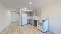 Located at 4700 Dixie Hwy NE in Palm Bay, Florida, this single-floor unit presents a clean, contemporary interior with a cohesive neutral palette throughout. Light wood-style luxury vinyl flooring runs continuously across the space, contributing to a bright and open atmosphere.

The kitchen is efficiently designed with gray flat-front cabinetry on both upper and lower levels, light countertops, a single-basin sink, a white refrigerator, and a white electric range. Recessed lighting illuminates the workspace, and a wall-mounted mini-split unit provides climate control for the main living area.

Adjacent to the kitchen, the open living area benefits from a window that frames views of mature Florida vegetation, allowing natural light to fill the space. The entry door is positioned nearby, offering a practical layout that connects the main living and kitchen zones seamlessly.

The bathroom is finished in the same light tones as the rest of the unit, featuring tile-style flooring, a vanity with cabinet storage and a large mirror with overhead lighting, and a full toilet. The overall design across the unit is clean and minimalist, with white walls and simple fixtures that create a fresh, move-in-ready environment suitable for comfortable everyday living.
