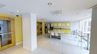 This single-floor space makes a striking visual impression through its bold use of color and contemporary design. The open-concept layout centers around a vibrant yellow kitchen, where floor-to-ceiling cabinetry with frosted panel insets lines the wall, providing generous storage. The kitchen is equipped with stainless steel appliances, including a side-by-side refrigerator and microwave, complemented by a sleek gray countertop that extends the full length of the cabinetry. A large central island or peninsula serves as both a workspace and casual dining surface, accompanied by modern white molded chairs that add to the space's clean aesthetic. Recessed lighting is installed throughout the ceiling, ensuring consistent and even illumination across the entire area. The flooring consists of large-format white ceramic tiles that reflect light and enhance the sense of openness. Structural white columns punctuate the space without interrupting the overall flow. Toward the rear, a corridor leads to what appears to be an elevator or secondary room, flanked by warm-toned glass paneling that introduces a subtle contrast to the otherwise white and yellow palette. The overall ambiance is clean, functional, and energetic, well-suited for a communal or shared-use environment.