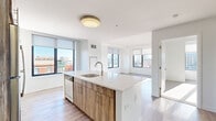 Located in the heart of downtown Stamford, CT, this well-appointed single-floor unit offers a thoughtfully designed layout across four distinct areas. The open-concept living and kitchen space serves as the centerpiece, featuring a large island with a quartz countertop, an undermount sink with a brushed gold faucet, and stainless steel appliances including a refrigerator, range, and microwave. Cabinetry combines white uppers with warm wood-toned lowers, complemented by a white tile backsplash. Light wood-style flooring flows throughout the main living areas, enhancing the bright, airy atmosphere created by multiple large, black-framed windows that frame sweeping views of the Stamford skyline. The living area is generously proportioned, bathed in natural light from corner and wall-facing windows with roller shades. The bedroom features plush carpeting and its own city-view window, offering a quiet retreat. The bathroom is finished with clean white subway tile surrounding a soaking tub and shower combination, brushed nickel fixtures, and neutral tile flooring. Clean white walls and modern flush-mount lighting throughout give the space a crisp, contemporary feel well-suited to urban living.