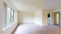 This single-floor residence offers a thoughtfully laid-out floor plan with generously sized living spaces throughout. The main living area is broad and open, featuring soft carpeting, neutral cream-toned walls, and a textured ceiling. Large windows along the exterior walls allow natural light to fill the room, while a sliding glass door in one of the rooms opens directly to a lush, tree-lined outdoor area, providing a pleasant connection to nature.

The kitchen is functionally designed with rich, dark wood cabinetry offering ample storage, light-colored countertops, and a suite of white appliances including a refrigerator, electric range, over-the-range microwave, and a double sink. Light vinyl flooring grounds the space and contrasts nicely with the warm wood tones of the cabinetry.

The bedrooms are carpeted and well-proportioned, each featuring windows that bring in natural light and views of the surrounding greenery. The rooms provide a calm, neutral backdrop suitable for a variety of layouts and uses.

The bathroom is finished with classic mosaic tile flooring in white and green, complemented by sage-green wall tile that extends partway up the walls. A single-sink vanity with white cabinetry, a toilet, and a bathtub with a tiled surround complete the space. A louvered cabinet door adds a traditional touch to the overall design.