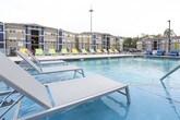 This single-floor community amenity space is designed with a contemporary aesthetic, offering residents a well-rounded social and recreational environment. The interior opens into a vibrant game room area, where light wood-look flooring, vaulted ceilings, and bold pop-art wall graphics create an energetic atmosphere. A full-size ping-pong table serves as a central focal point, complemented by casual seating with colorful accent pillows. Adjacent to the game room, a billiards table occupies a stylishly separated zone, framed by dramatic floor-to-ceiling draped curtains that add texture and visual depth. A striking marble-clad accent wall houses a sleek linear fireplace with a wall-mounted television above, providing an elegant lounging area that balances the playful recreational elements. Large glass-paned double doors connect the interior to the outdoor pool deck, allowing abundant natural light to fill the space and providing seamless indoor-outdoor flow. Outside, a well-appointed pool deck features a row of modern lounge chairs in cheerful green and yellow tones arranged alongside the pool, enclosed by a wrought iron perimeter fence. Together, these spaces form a cohesive, amenity-rich environment tailored for both relaxation and social activity.