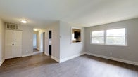 This single-story space offers a clean, functional layout designed for comfortable everyday living. The main living area is finished with smooth hardwood-style flooring in a warm gray-toned hue, complemented by crisp white baseboards and neutral light gray walls that create a bright, cohesive atmosphere throughout. A large window in the living area allows natural light to filter in, offering a view of the surrounding greenery outside.

A defining architectural feature is the pass-through window cut into the interior wall, which connects the living area to the kitchen. This opening provides a sense of visual connectivity between the two spaces while maintaining a distinct separation. The kitchen, visible through the pass-through, features dark cabinetry that provides a striking contrast to the lighter tones of the surrounding walls.

A hallway extending from the living area leads further into the home, with additional rooms accessible from it. The hallway is well-lit with overhead lighting and continues the same flooring material for a consistent aesthetic. Overall, the space presents a straightforward, move-in-ready environment with a neutral palette well-suited for a variety of interior styles.