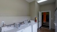 This single-floor laundry facility is designed with practicality and functionality in mind. The room features a row of white coin-operated top-loading washing machines lined along one wall, providing ample capacity for multiple users. The walls are finished in a neutral taupe tone, giving the space a clean and understated aesthetic that complements the white appliances. Tile flooring runs throughout the room, offering a durable and easy-to-maintain surface well-suited for a high-traffic laundry environment. The ceiling is elevated, contributing to an open and airy feel within the space. Overhead fluorescent lighting ensures the room is well-lit, supporting visibility and ease of use. A fire extinguisher is mounted on the wall, reflecting attention to safety standards within the facility. An electrical outlet is accessible along the wall, adding utility to the space. A white paneled door leads to an adjoining area, glimpses of which reveal an orange accent wall, suggesting additional connected rooms or common areas. Overall, the laundry room is a well-maintained, efficiently organized shared facility that prioritizes user convenience and safety.