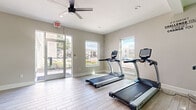 This single-floor fitness center is thoughtfully designed to accommodate a wide range of workout preferences in a clean, modern environment. The space features light wood-style flooring throughout, neutral gray walls, and white trim that create a bright, welcoming atmosphere. Large windows and glass entry doors flood the interior with natural light while offering views of the surrounding outdoor landscape.

The main cardio area is generously sized, housing multiple treadmills and elliptical machines from the True brand, complemented by overhead ceiling fans that promote airflow during intense workouts. Adjacent to the cardio zone is a dedicated cycling area equipped with several stationary spin bikes and a decorative indoor tree that adds a lively, energetic touch to the space. Motivational wall decals, including phrases like "You Can – End of Story" and "Student Challenge You, Change You," reinforce an encouraging workout environment.

A separate strength and functional training room features wall-mounted foam rollers, a medicine ball rack, dumbbells, a weight bench, a pull-up and cable machine, and additional resistance equipment. Recessed lighting in this zone ensures it remains well-lit regardless of natural light availability.

Throughout the facility, multiple entry and exit points provide convenient access, and the overall layout supports a smooth flow between workout zones.