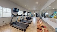 This fitness center offers a spacious, single-floor layout designed to accommodate a comprehensive range of workout activities. The interior is finished with light wood-look flooring throughout, white walls, and recessed lighting that creates a clean, well-lit environment. Natural light filters in through multiple windows along the perimeter, enhancing the open and airy feel of the space.

The cardio zone features a row of treadmills and an elliptical trainer, each positioned in front of wall-mounted flat-screen televisions for entertainment during workouts. A full-length mirror spans one wall, visually expanding the room while allowing users to monitor their form. A dumbbell rack stocked with a wide range of free weights anchors the strength training area, complemented by a multi-function cable machine and a workout bench nearby.

A dedicated functional training area includes stability balls, medicine balls, resistance equipment, and foam rollers neatly organized on a storage rack. A stationary bike rounds out the cardio offerings. The entry area includes a water cooler station, a bulletin board, and a mini-split HVAC unit, ensuring comfort year-round. A colorful area rug adds a warm visual accent throughout the main floor space.